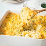 A wooden spoon lifts a serving of cheesy broccoli rice casserole from a white baking dish, revealing gooey melted cheese and fresh herbs on top.