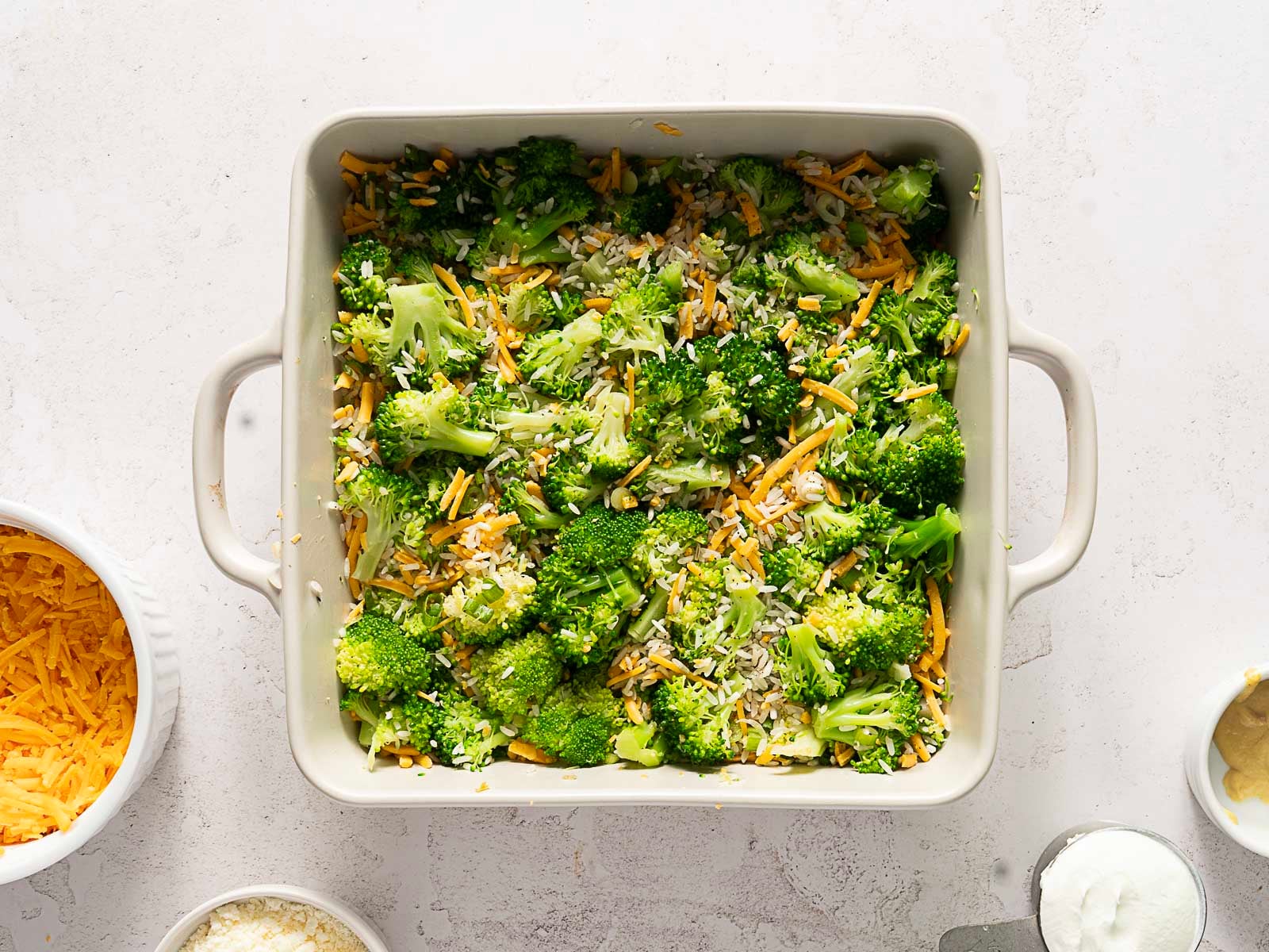 A white square baking dish filled with chopped broccoli, shredded cheese, and grains sits on a white surface, surrounded by bowls of ingredients-perfect for making a delicious Cheesy Broccoli Rice Casserole.