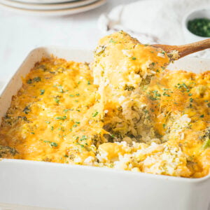 A wooden spoon lifts a serving of cheesy broccoli rice casserole from a white baking dish, revealing gooey melted cheese and fresh herbs on top.