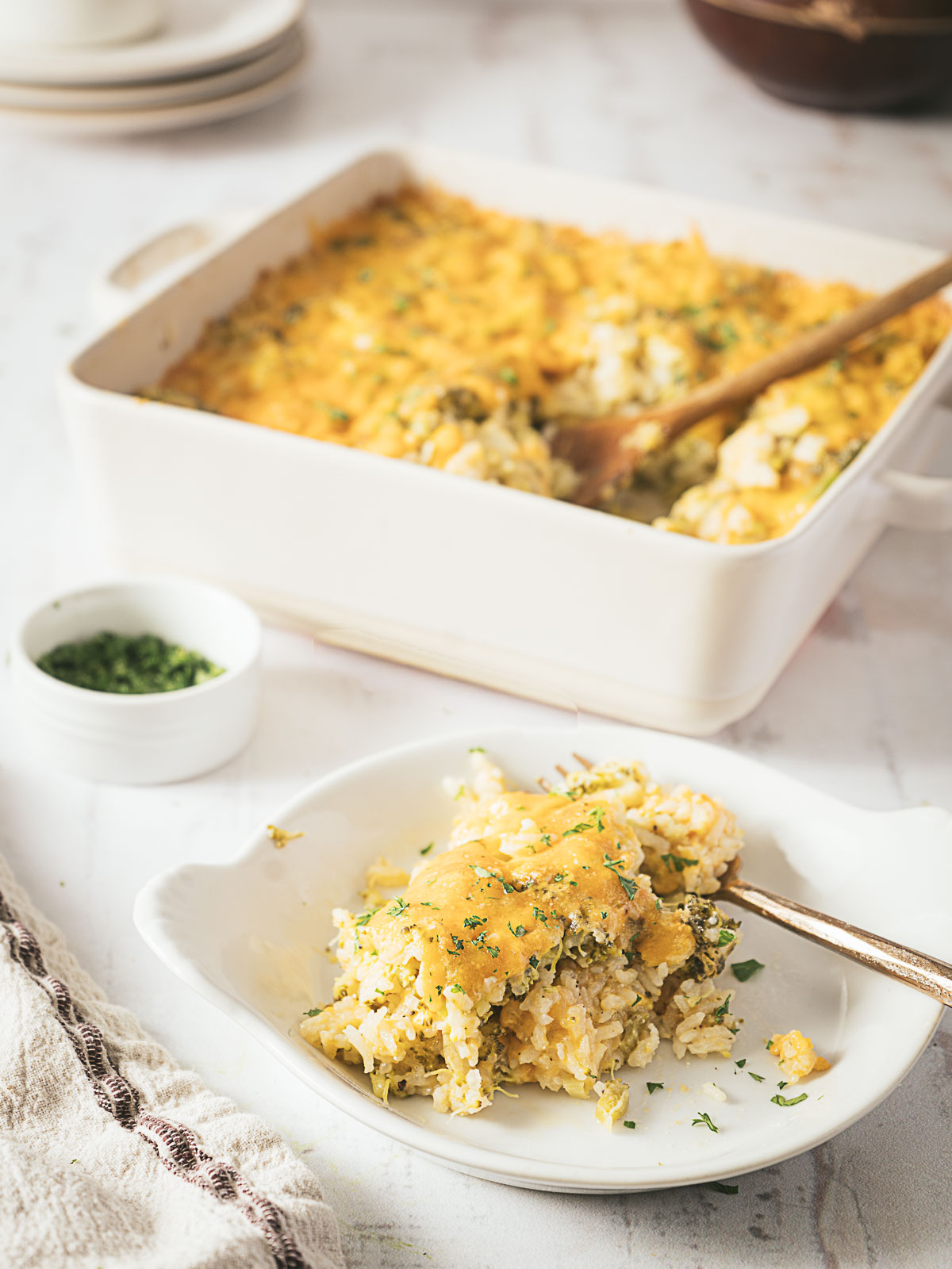 A serving of cheesy broccoli rice casserole on a plate with a fork, with the casserole dish and a small bowl of herbs in the background.