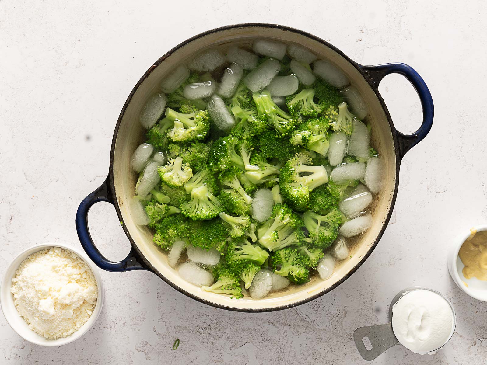 A pot of broccoli florets sits in ice water; nearby are bowls containing grated cheese, mustard, and a cup of yogurt-perfect ingredients for a cheesy broccoli rice casserole.