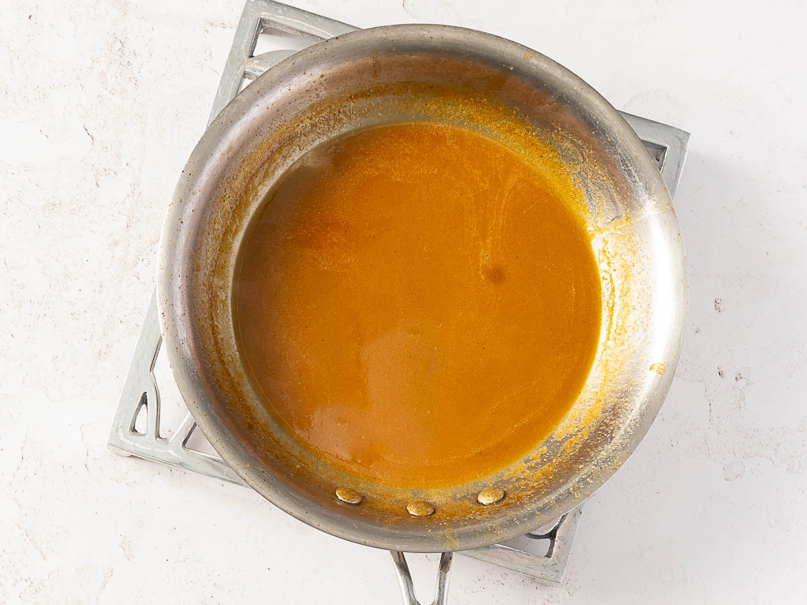 A stainless steel pan containing a small amount of brown pan sauce, perfect for drizzling over bundt pan chicken, sits on a metal trivet on a white surface.