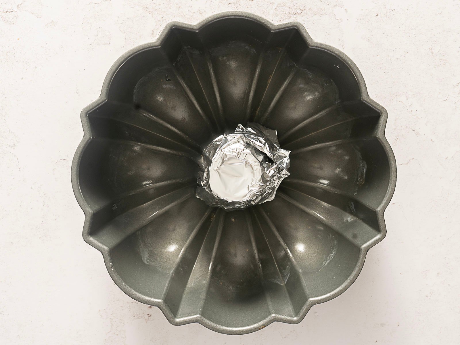 A Bundt cake pan, perfect for making bundt pan chicken, has its center hole filled and covered with crumpled aluminum foil, resting on a light-colored surface.