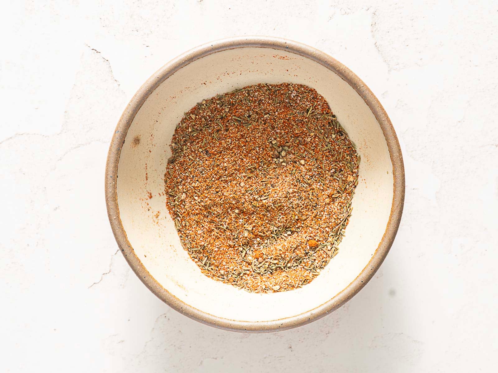 A bowl filled with a mixed blend of dried herbs and spices on a white surface, perfect for seasoning your next bundt pan chicken.