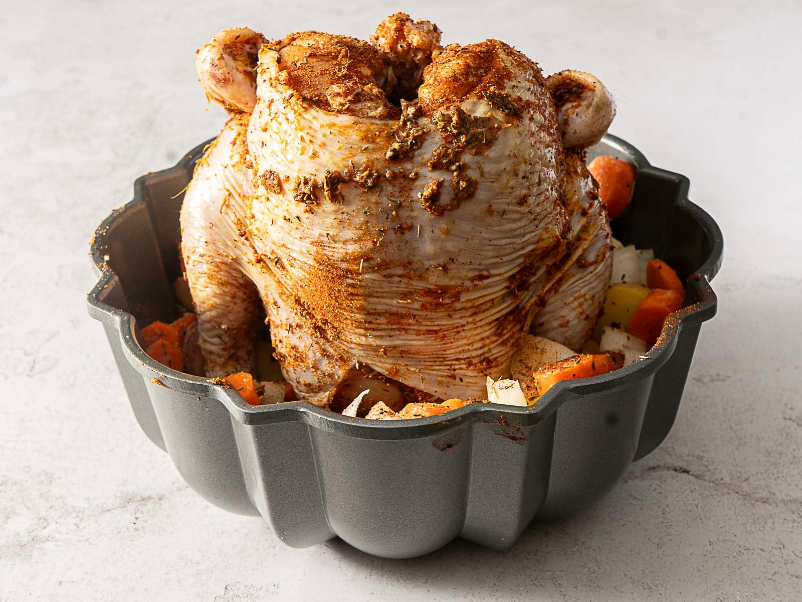 A seasoned bundt pan chicken sits upright in the pan, surrounded by chopped vegetables on a light-colored surface.