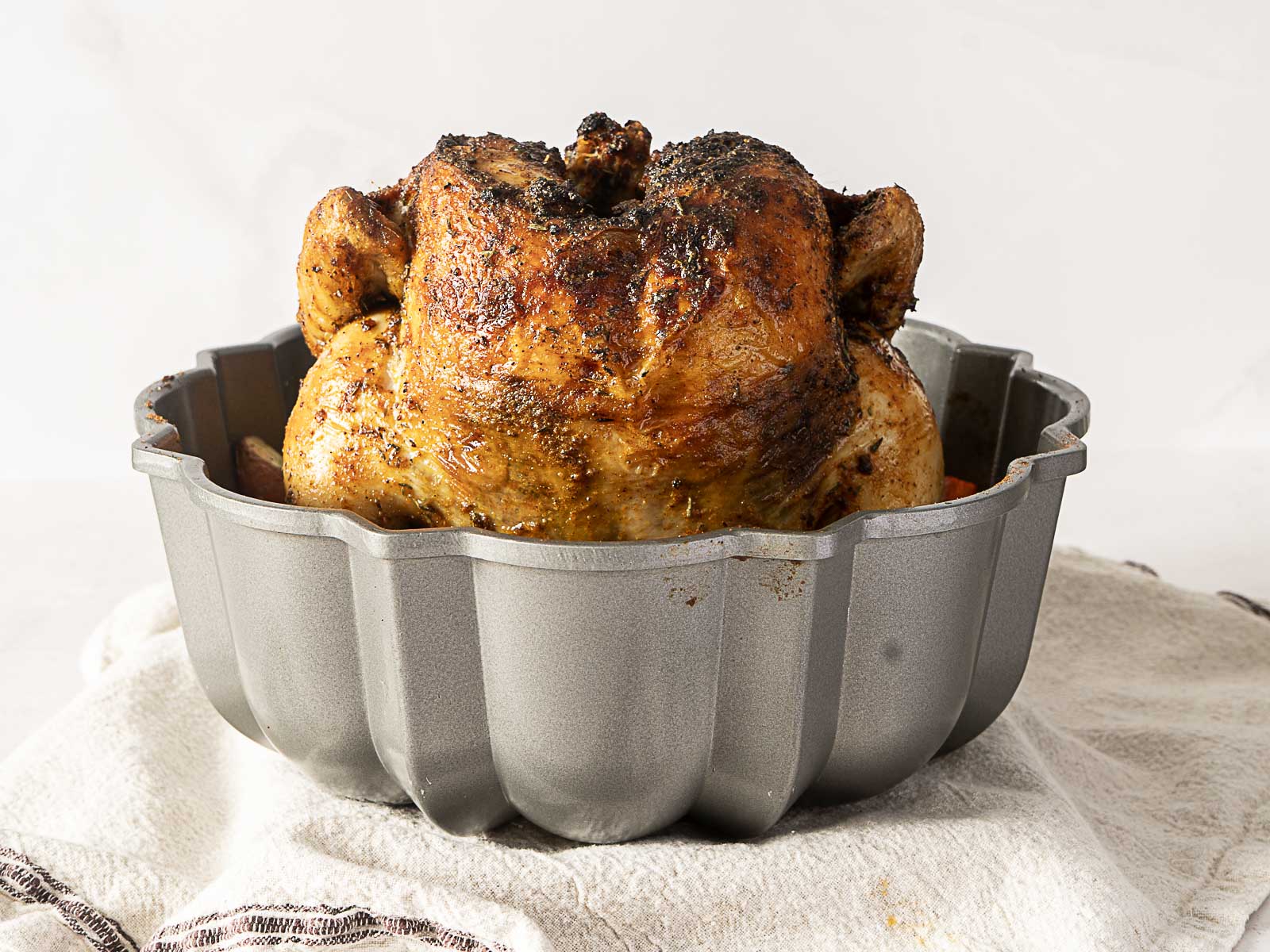 A whole roasted bundt pan chicken sits upright in a gray Bundt pan on a towel-covered surface.