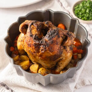 A whole roasted bundt pan chicken with browned skin sits surrounded by roasted potatoes and carrots, with a bowl of chopped herbs in the background.