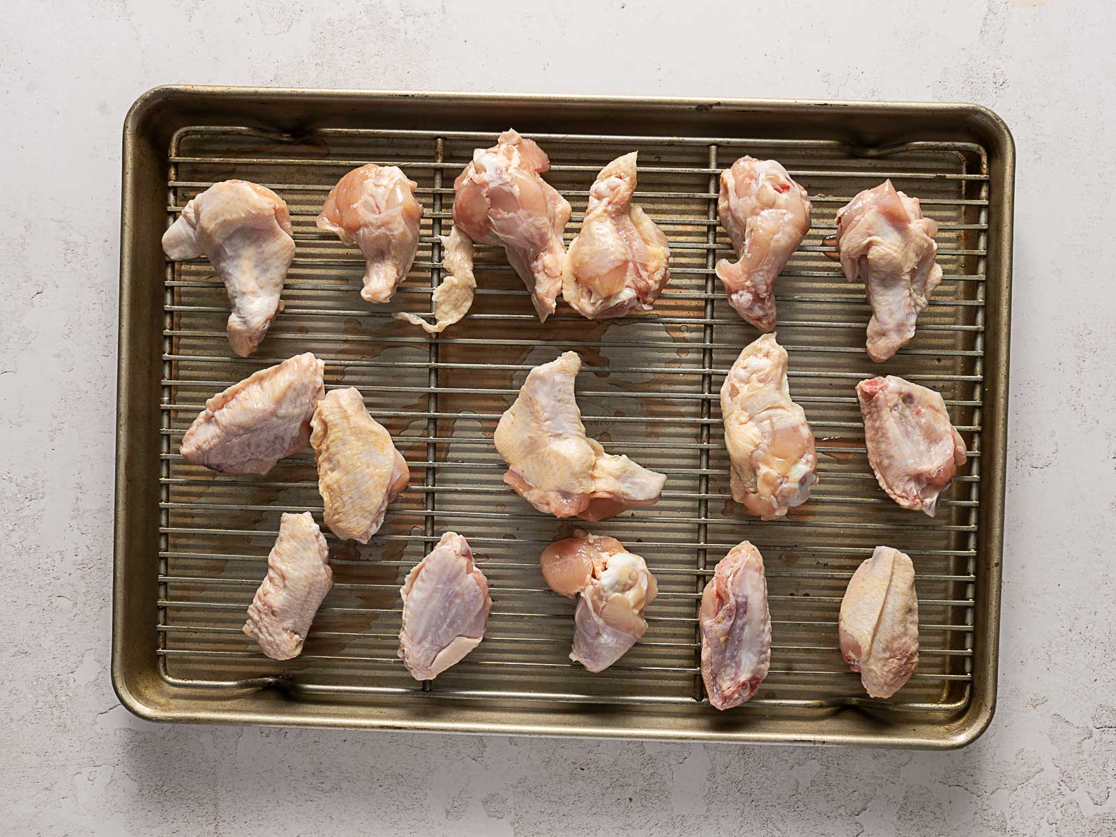 Raw chicken wings, soon to become Hot Honey Buffalo Wings, are arranged on a wire rack set over a baking sheet, ready for cooking. The baking sheet rests on a light-colored countertop.