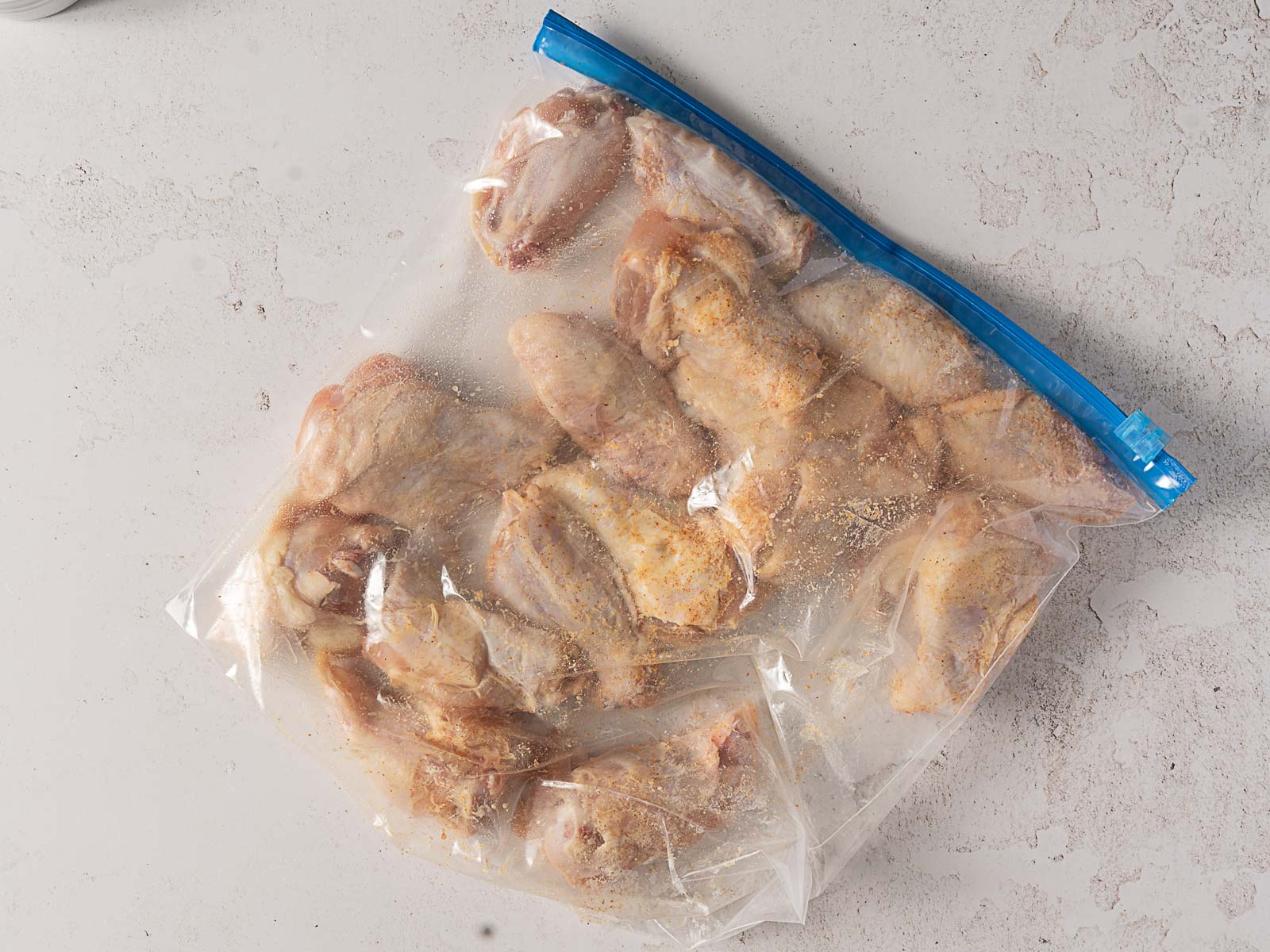 A clear resealable plastic bag filled with raw Hot Honey Buffalo Wings sits on a light-colored surface. The chicken wings are coated in seasoning, partially sealed in the bag, and evenly distributed inside.