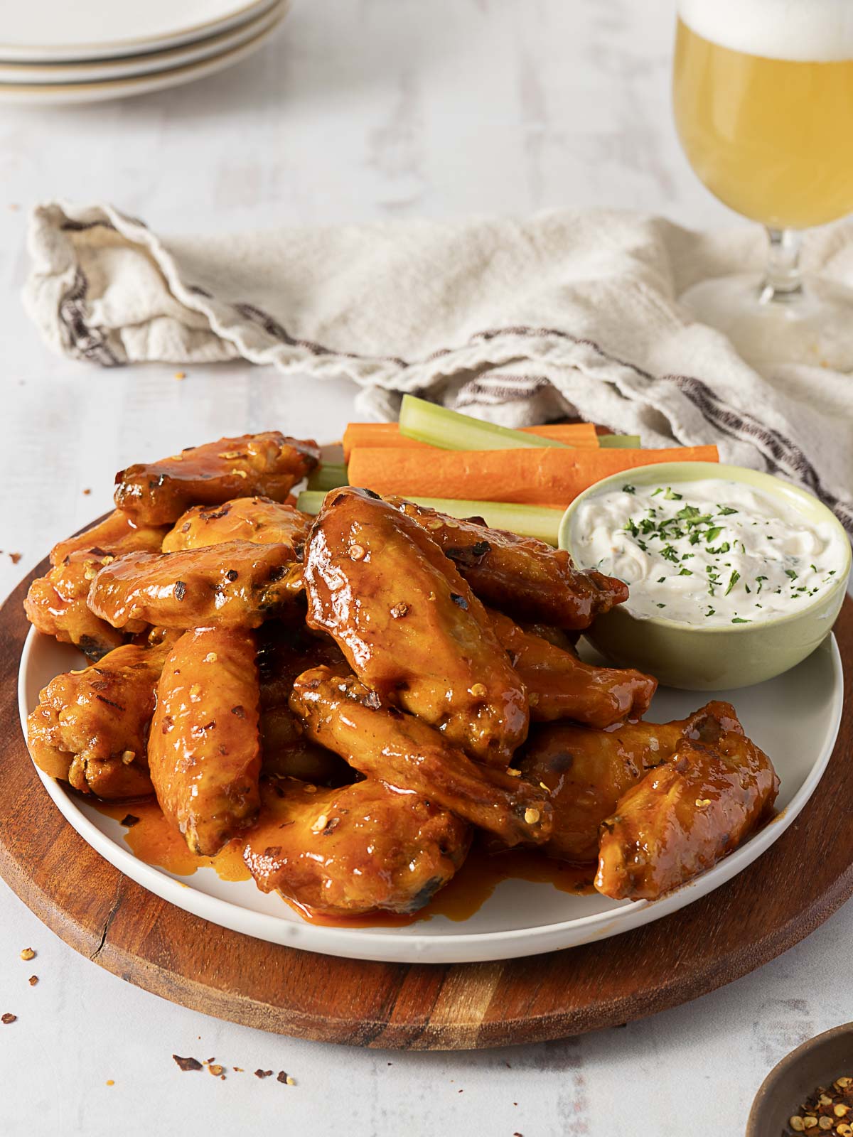 A plate of saucy Hot Honey Buffalo Wings is served with carrot and celery sticks, a bowl of ranch dip, and a glass of beer on a wooden board, with a napkin and plates in the background.