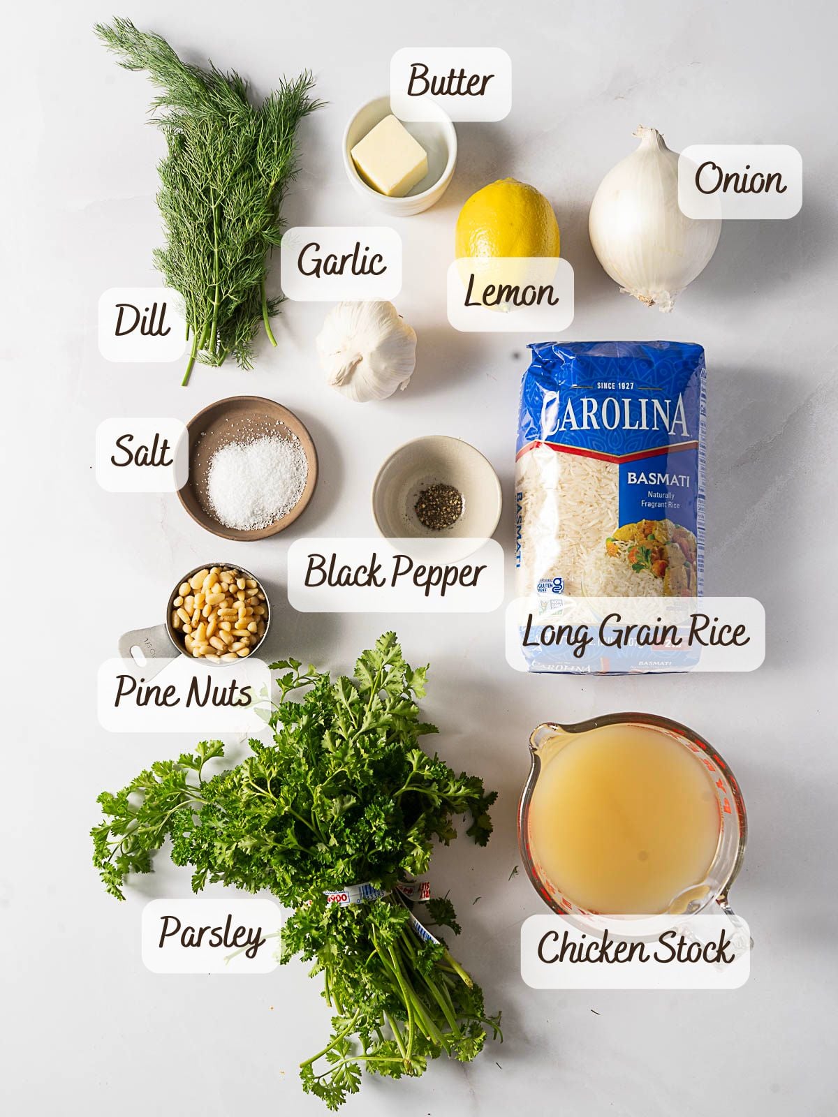 Assorted ingredients labeled for a Greek Rice Pilaf recipe, including dill, parsley, pine nuts, salt, black pepper, butter, garlic, lemon, onion, basmati rice, and chicken stock on a white surface.