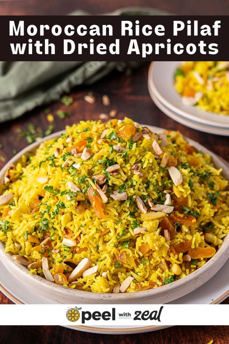 A plate of Moroccan rice pilaf topped with dried apricots, chopped herbs, and slivered almonds, with another plate and a green cloth in the background.
