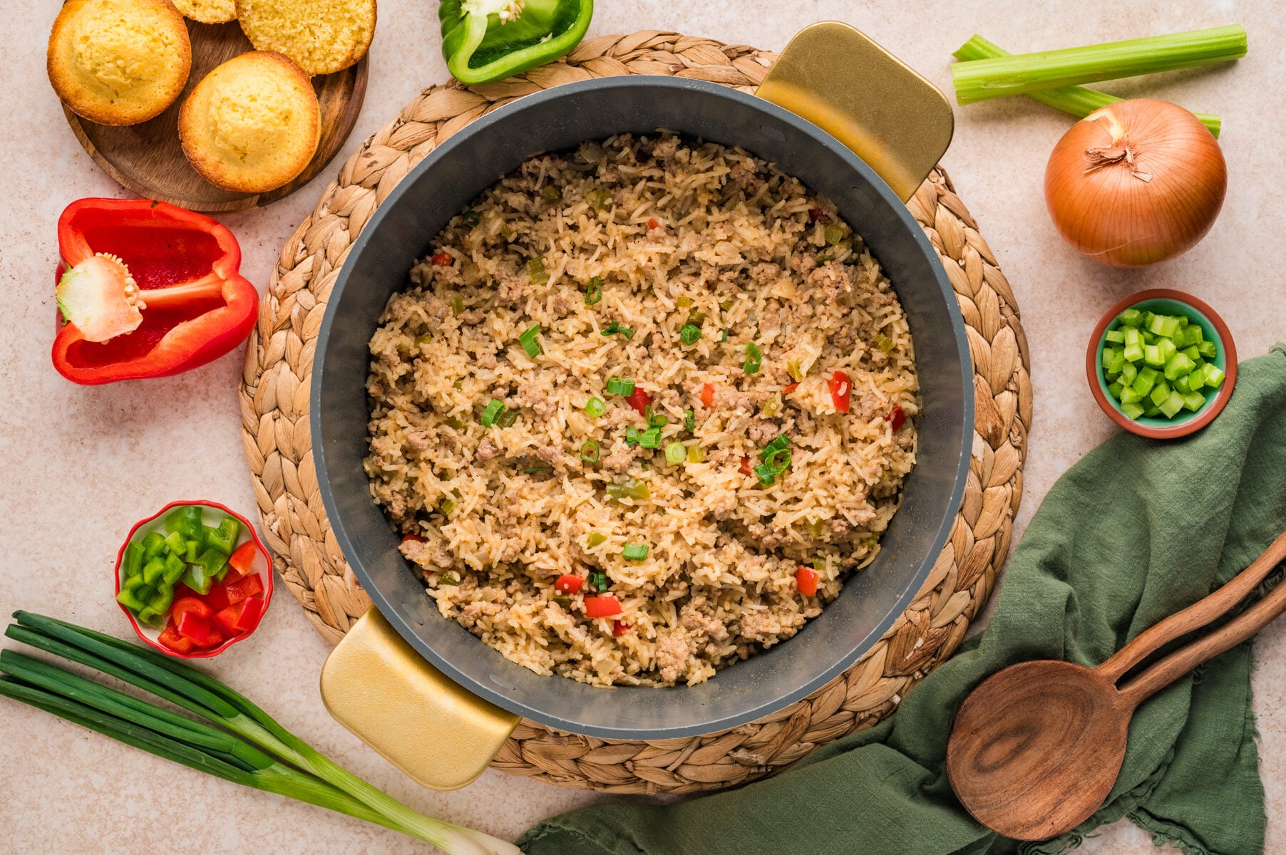 A pot of Cajun Dirty Rice with ground meat and diced vegetables, surrounded by fresh ingredients, cornbread muffins, and wooden utensils on a woven mat.