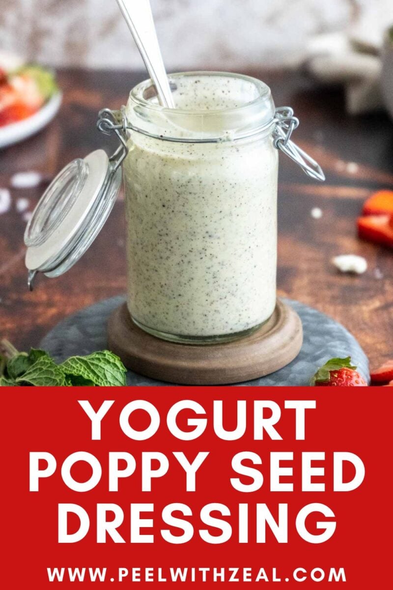 A glass jar filled with creamy yogurt poppy seed dressing, with a spoon inside, sits on a wooden coaster.
