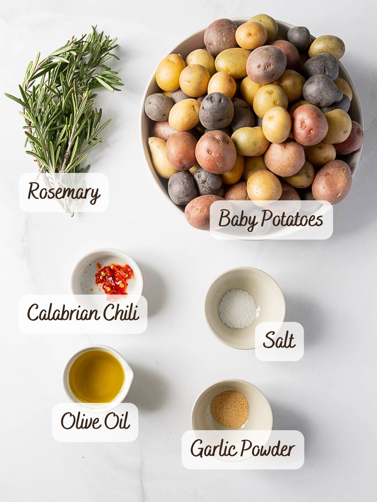A bowl of mixed baby potatoes, perfect for rosemary roasted potatoes, sits beside fresh rosemary and small dishes of chopped Calabrian chili, salt, olive oil, and garlic powder on a white surface.