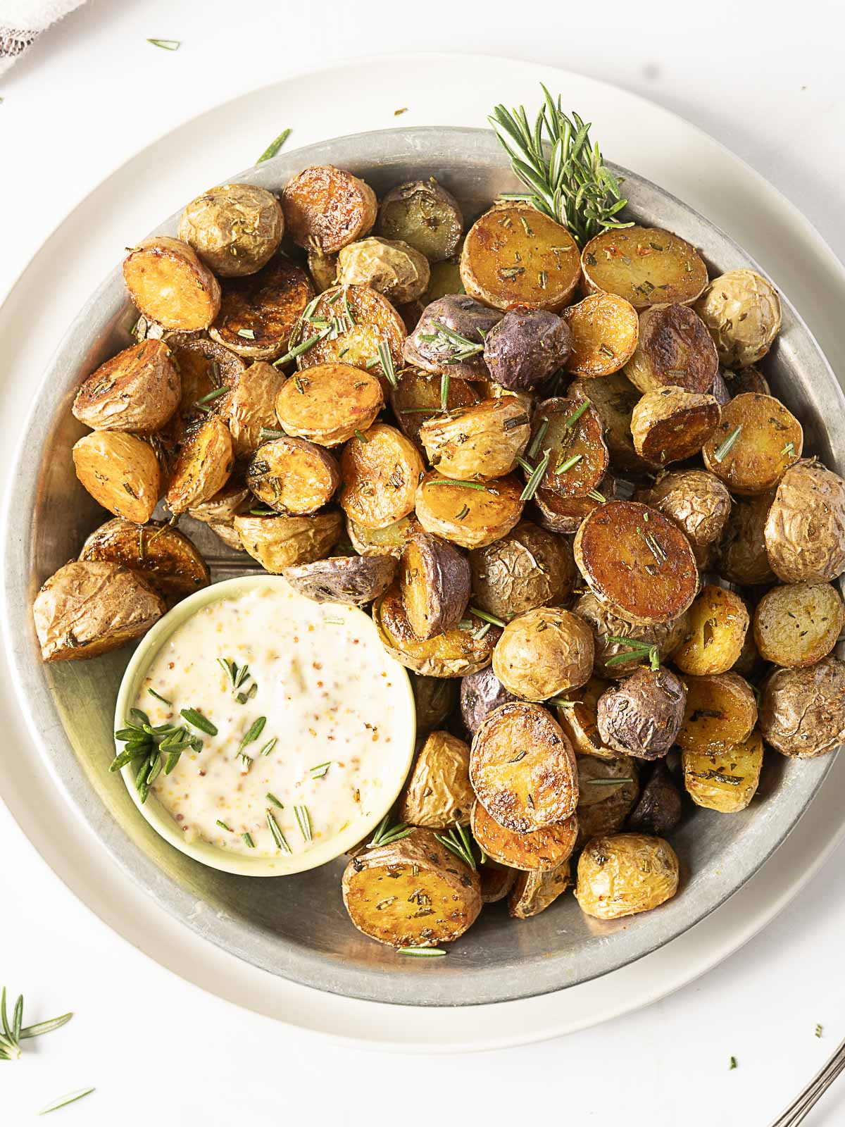 A plate of crispy baby potatoes, perfectly roasted and garnished with rosemary, is served alongside a small bowl of creamy dipping sauce.