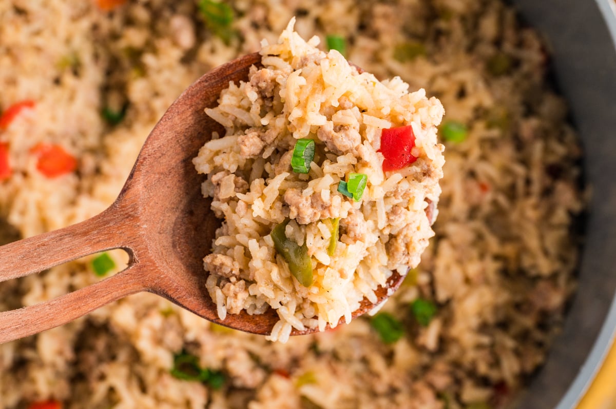 A wooden spoon holds a serving of Cajun Dirty Rice&mdash;rice mixed with ground meat, diced red peppers, green onions, and green peppers&mdash;above a pan filled with the flavorful dish.