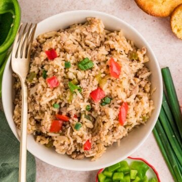 A bowl of Cajun Dirty Rice mixed with ground meat, diced red bell peppers, and chopped green onions, with a fork resting on the side. Green onions and bell pepper pieces are nearby.