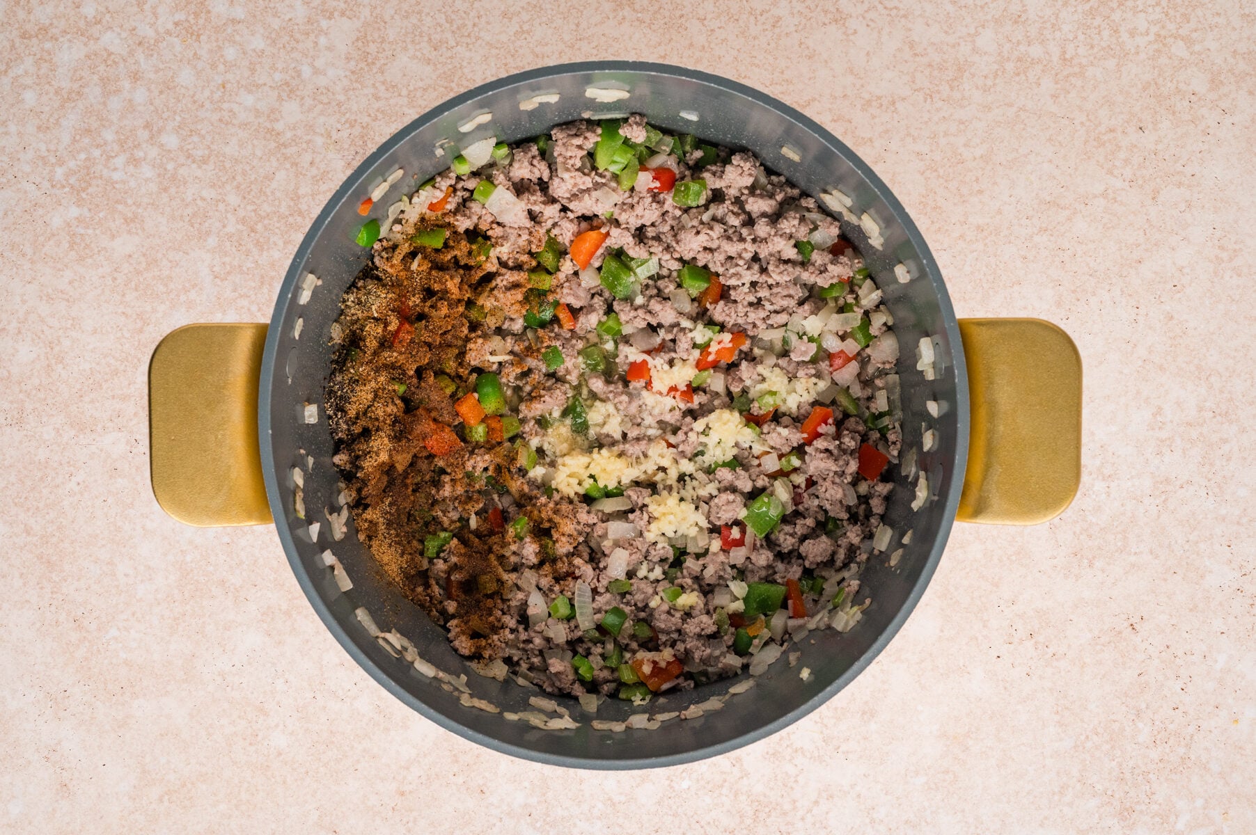 A pot containing cooked ground meat, chopped onions, green and red bell peppers, and various spices, viewed from above on a light-colored surface-classic ingredients for savory Cajun Dirty Rice.