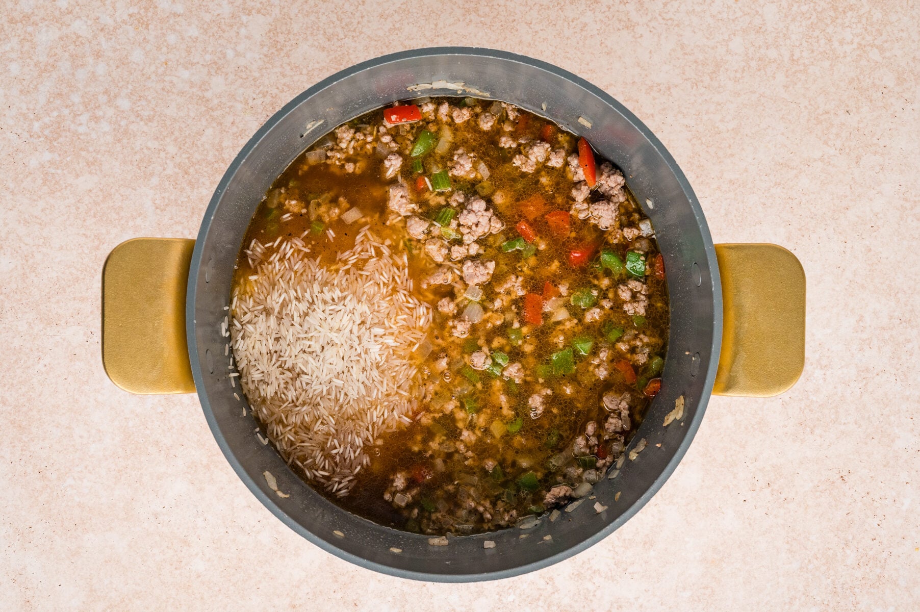 A pot containing uncooked rice, ground meat, chopped vegetables, and broth, viewed from above on a light-colored surface-perfect for making classic Cajun Dirty Rice.