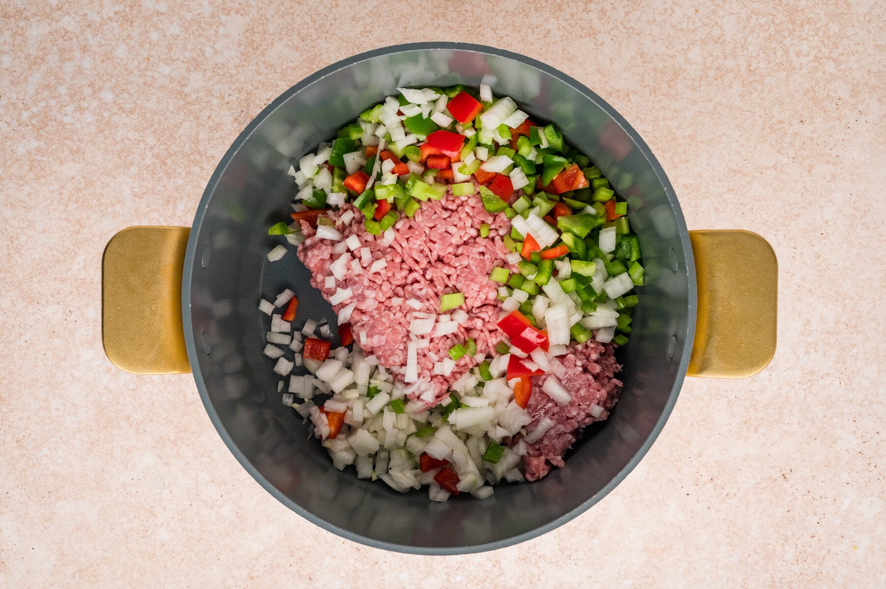 A pot containing chopped onions, green bell pepper, red bell pepper, and ground meat on a light countertop-classic ingredients for Cajun Dirty Rice.
