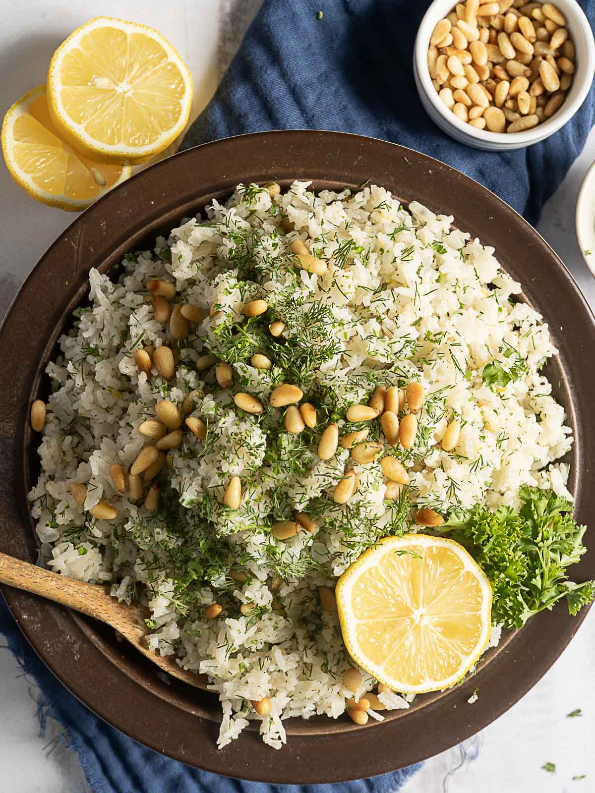 A bowl of Greek Rice Pilaf garnished with fresh dill, pine nuts, and parsley, with a lemon half and slices on the side. A wooden spoon rests in the bowl.