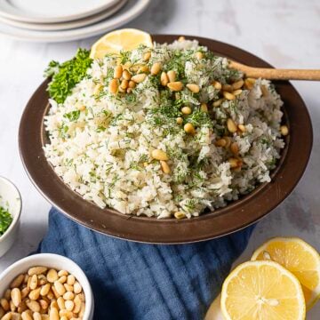 A bowl of Greek Rice Pilaf garnished with fresh herbs and pine nuts, served with lemon slices and parsley, with extra pine nuts and chopped herbs in small bowls nearby.