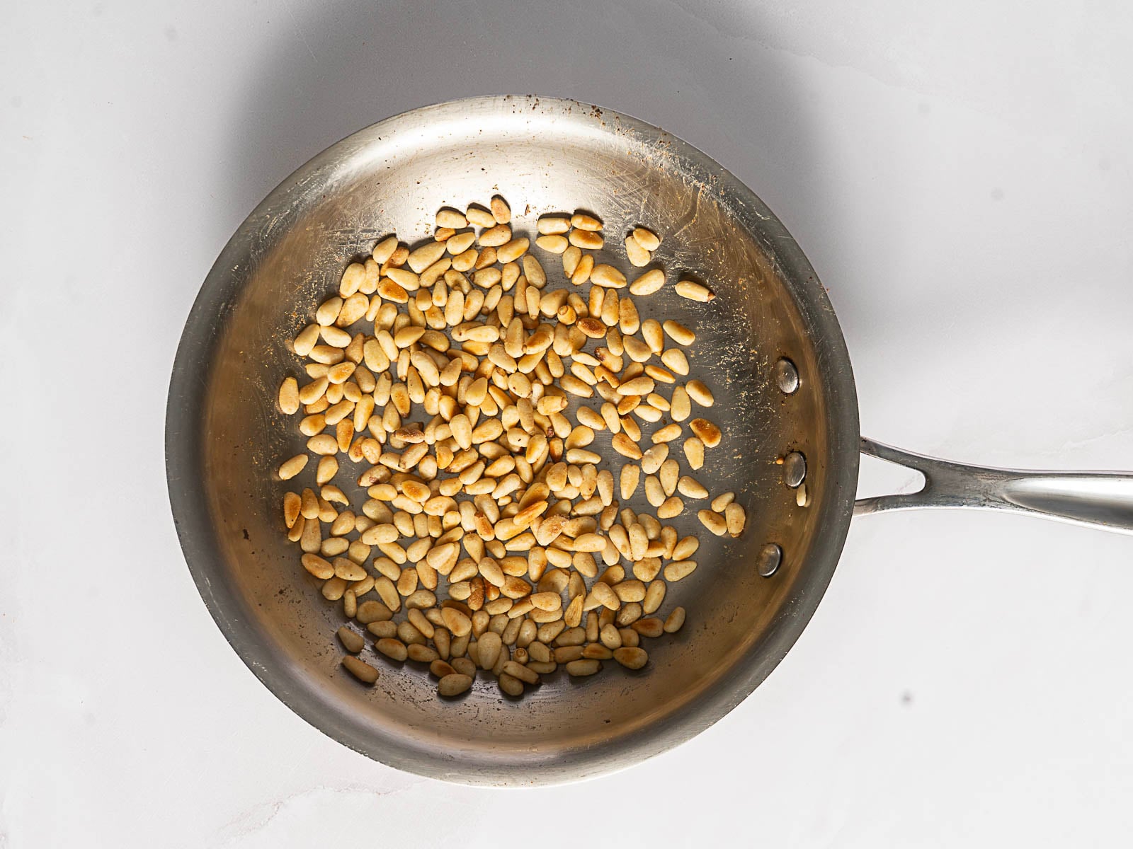 A stainless steel frying pan containing toasted pine nuts, perfect for adding to a classic Greek Rice Pilaf, is placed on a white surface.