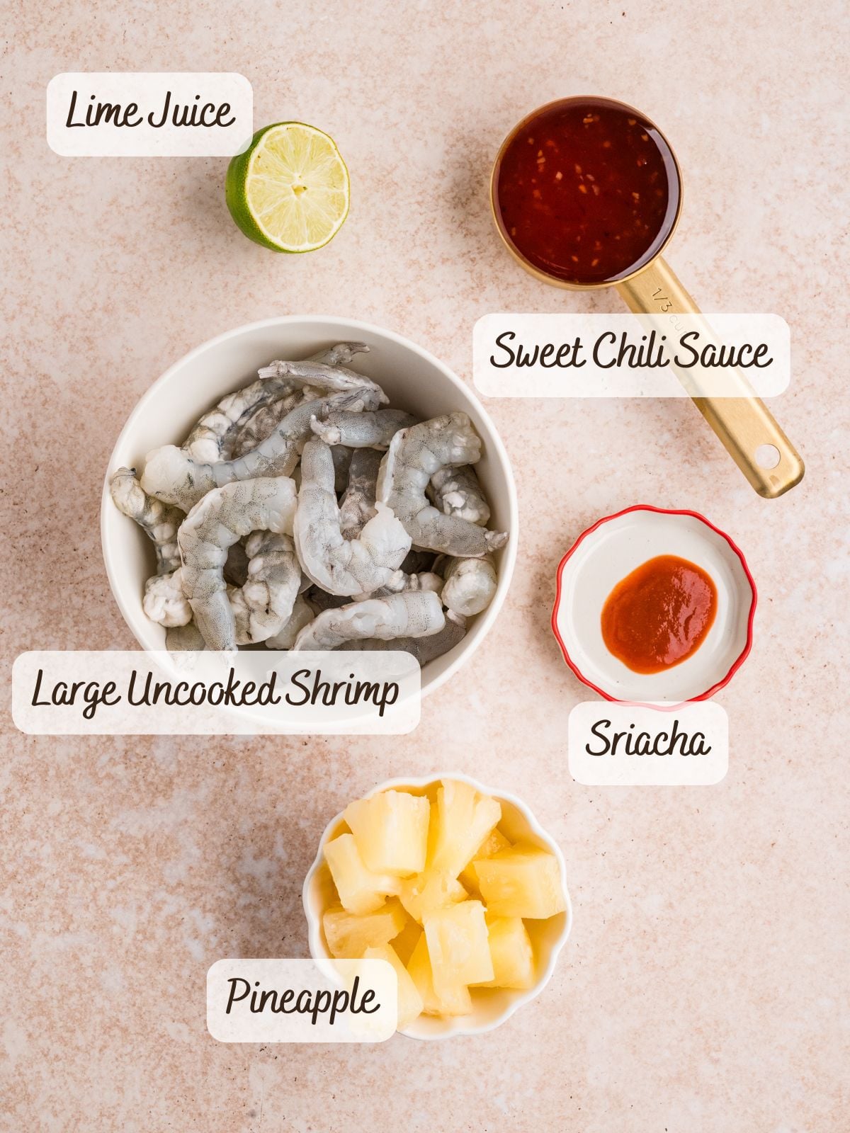 A bowl of raw shrimp, a cup of sweet chili sauce, a small dish of sriracha, lime half, and pineapple chunks are arranged on a countertop-perfect ingredients for tasty Shrimp Pineapple Skewers. Labels identify each item.