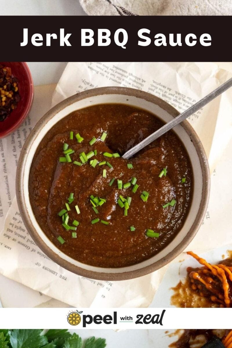 A bowl of jerk BBQ sauce garnished with chopped chives, with a spoon inside, placed on a paper with some sauce remnants and herbs nearby.