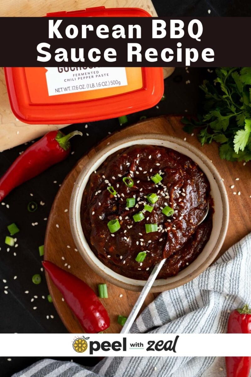 A bowl of Korean BBQ sauce garnished with sesame seeds and chopped green onions, surrounded by red chili peppers, a container of chili paste, and fresh cilantro on a black surface.