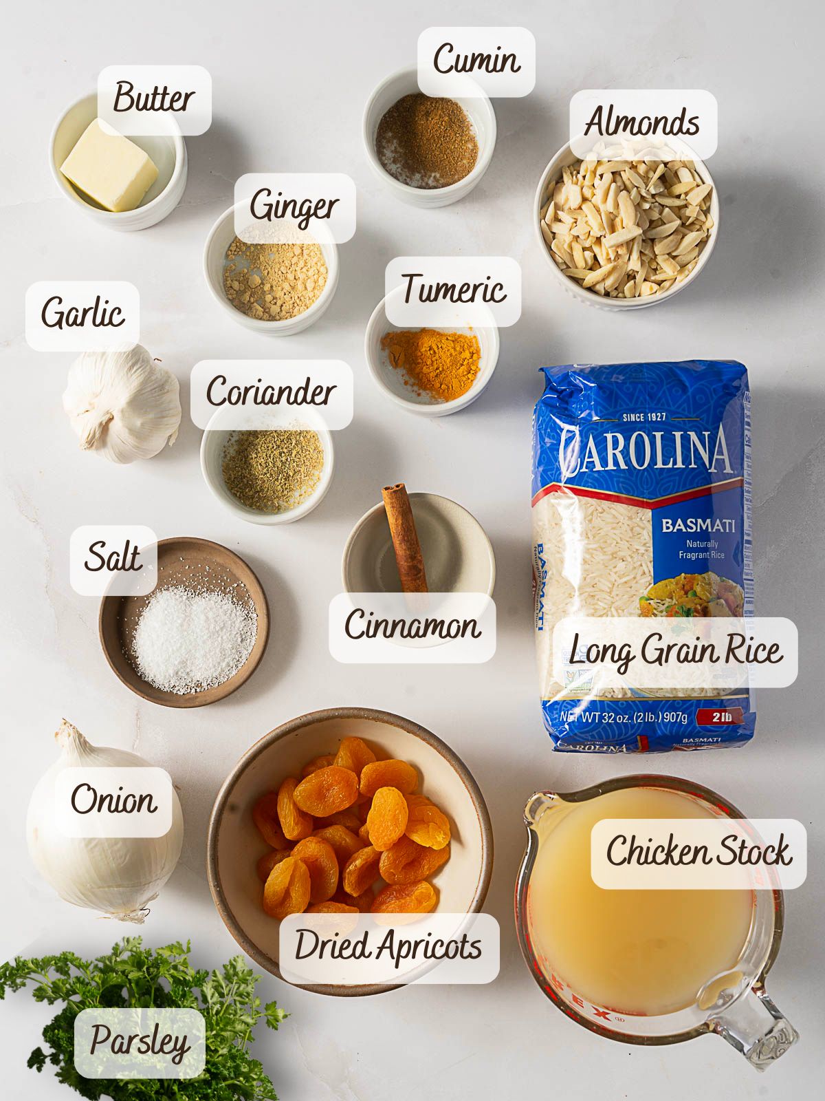 Ingredients for Moroccan Rice Pilaf laid out, including butter, cumin, ginger, almonds, turmeric, garlic, coriander, salt, cinnamon, long grain rice, onion, dried apricots, chicken stock, and parsley.