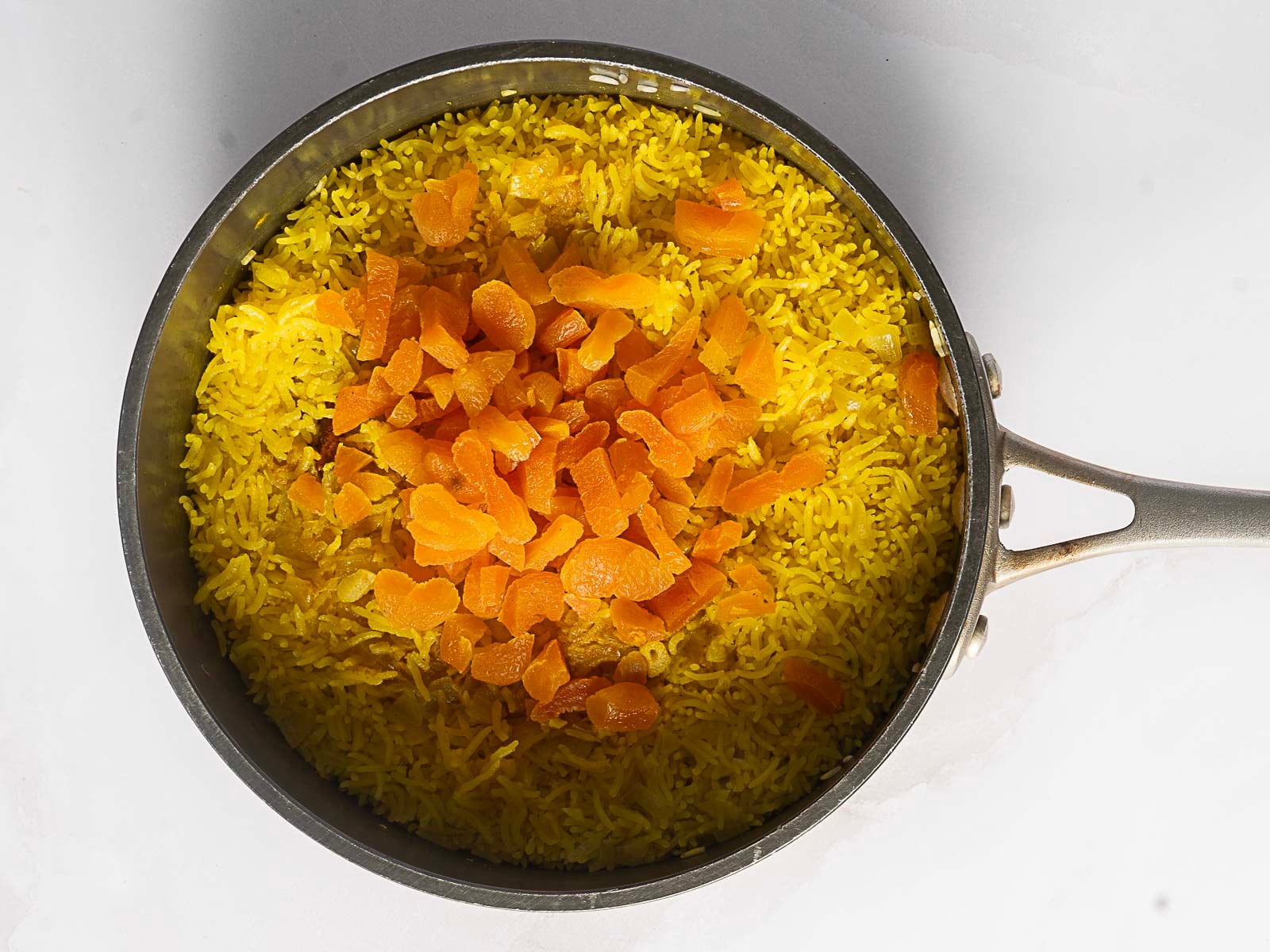 A saucepan filled with vibrant Moroccan Rice Pilaf, the yellow rice topped with a pile of chopped dried apricots, sits on a white surface.