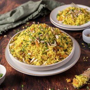 A plate of Moroccan Rice pilaf mixed with dried fruits and nuts, garnished with chopped herbs, sits on a wooden table with another serving in the background.