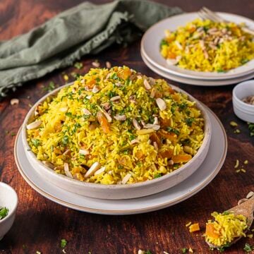 A plate of Moroccan Rice pilaf mixed with dried fruits and nuts, garnished with chopped herbs, sits on a wooden table with another serving in the background.
