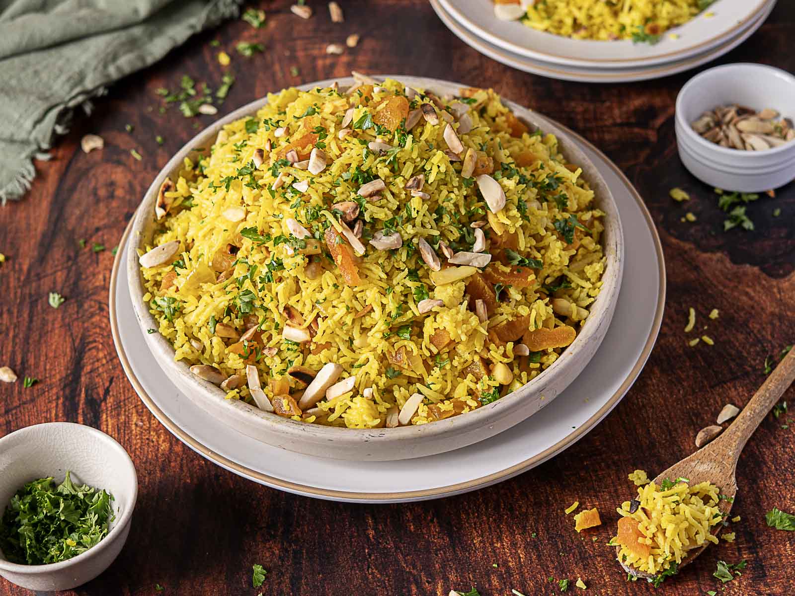 A plate of Moroccan Rice Pilaf garnished with chopped herbs and mixed nuts, with small bowls of herbs and nuts and a spoon on a wooden table.