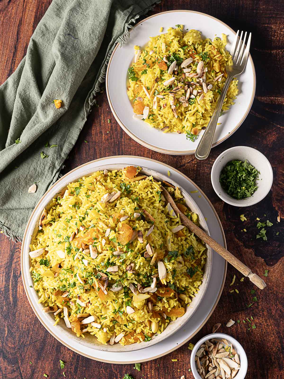 A large bowl and a plate filled with Moroccan Rice Pilaf, topped with slivered almonds and herbs, sit beside a small bowl of sliced almonds and a green napkin on a wooden table.