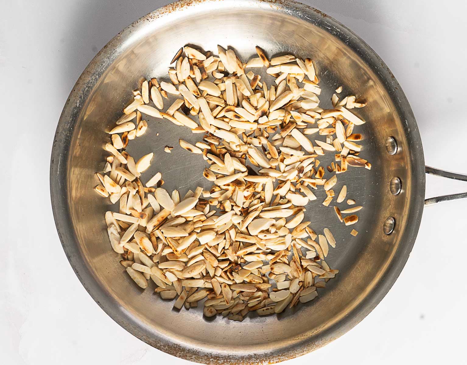 Slivered almonds being toasted in a stainless steel pan on a light-colored surface, perfect for adding to a Moroccan Rice Pilaf.