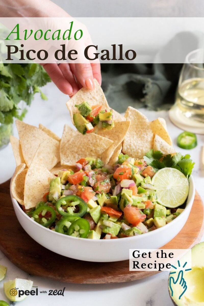 A hand dips a tortilla chip into a bowl of avocado pico de gallo garnished with lime and jalapeño slices, surrounded by chips. A glass of white wine is in the background.