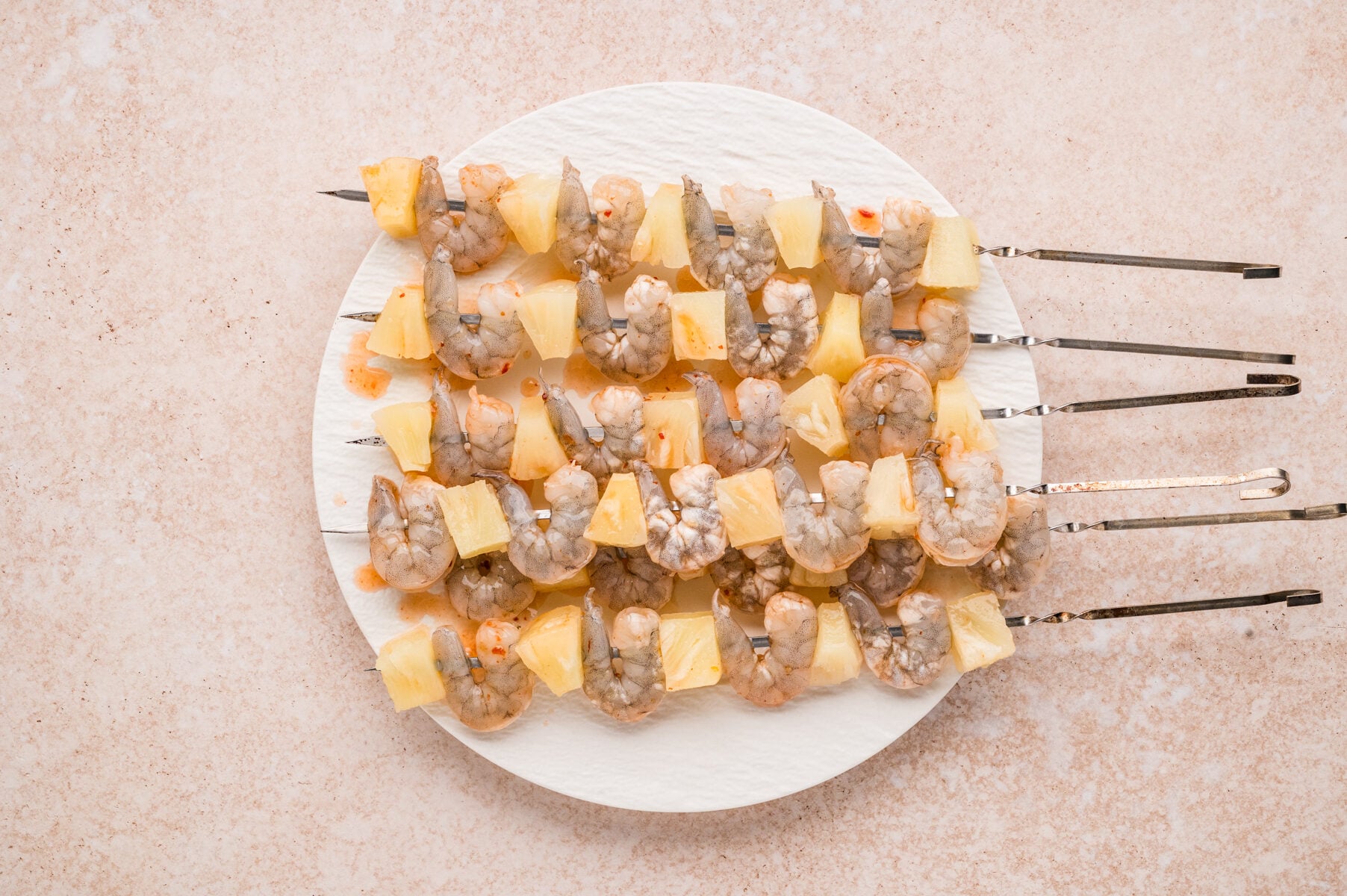 Shrimp Pineapple Skewers with alternating raw shrimp and pineapple pieces are arranged on a round white plate, placed on a light-colored surface.