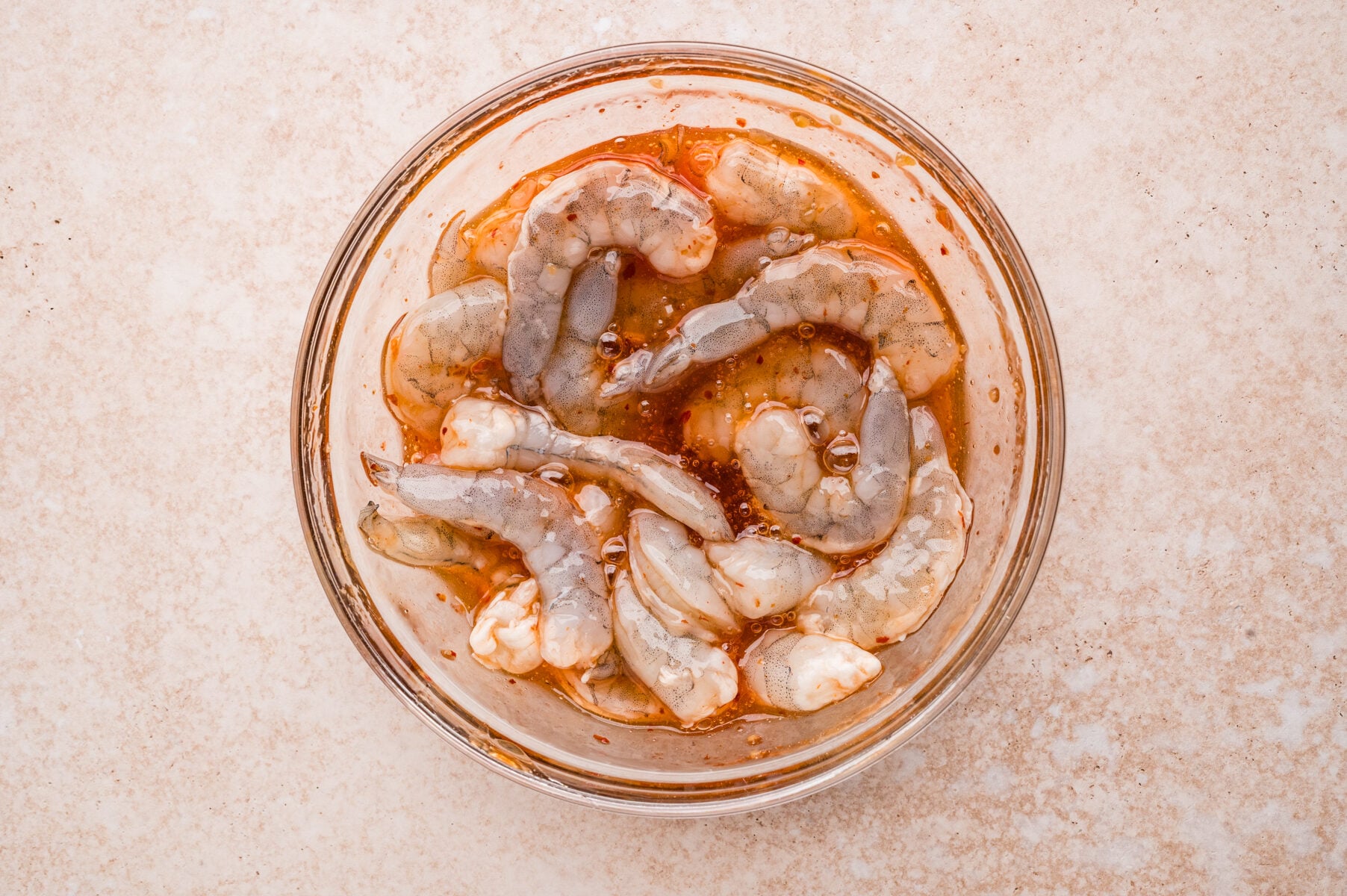 Raw shrimp marinating in a glass bowl with a reddish sauce, perfect for making flavorful Shrimp Pineapple Skewers, sits on a light-colored surface.