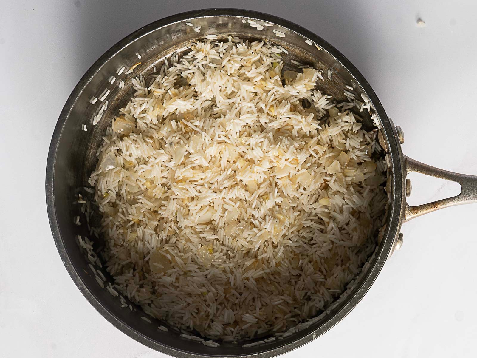 A metal saucepan containing uncooked Greek Rice Pilaf, with rice mixed and chopped onions, viewed from above on a white surface.