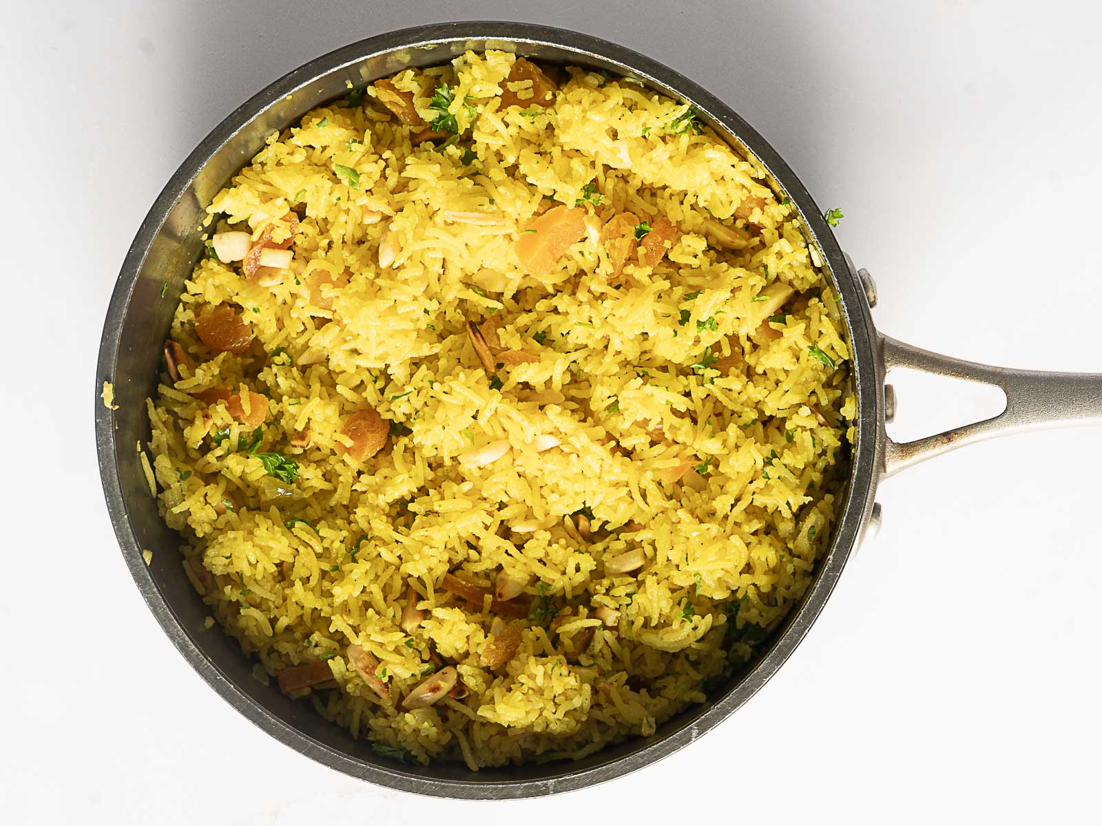 Overhead view of a pan filled with vibrant Moroccan Pilaf-yellow rice mixed with herbs and small pieces of nuts or vegetables-on a white background.