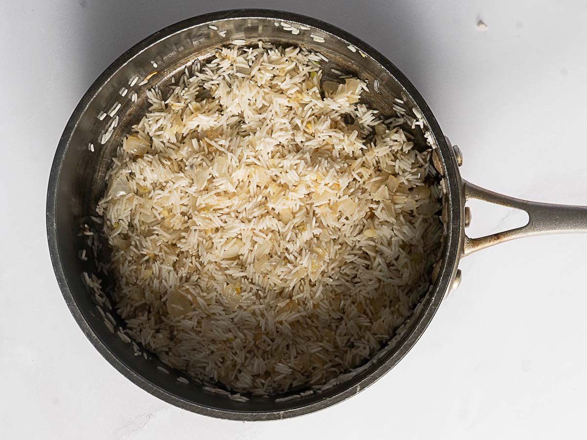 A metal pot containing uncooked Rice Pilaf mixed with saut&eacute;ed onions, viewed from above on a white surface.