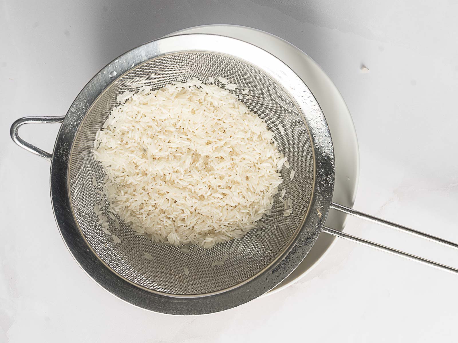 Uncooked white rice for Greek Rice Pilaf rests in a metal strainer positioned over a bowl on a white surface.