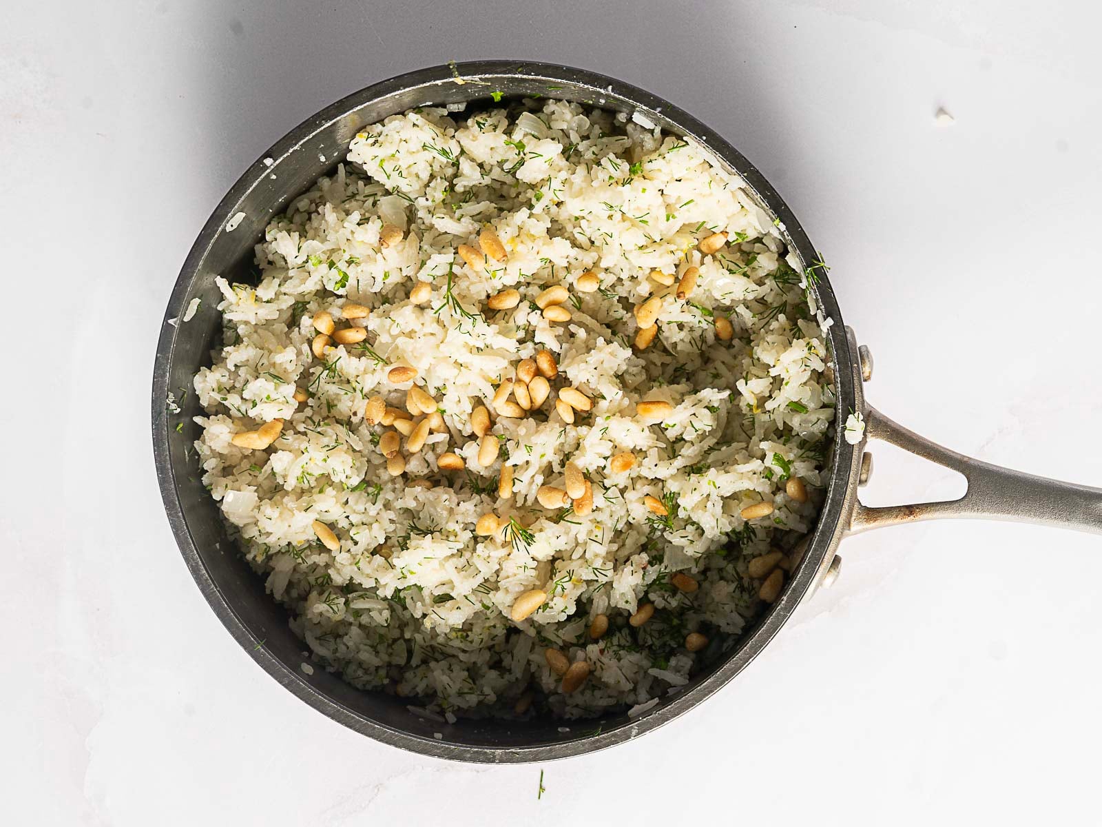 A saucepan filled with Greek Rice Pilaf-fluffy white rice mixed with herbs and topped with pine nuts-viewed from above on a light surface.