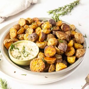 A plate of crispy baby potatoes garnished with herbs, served with a small bowl of creamy dipping sauce.