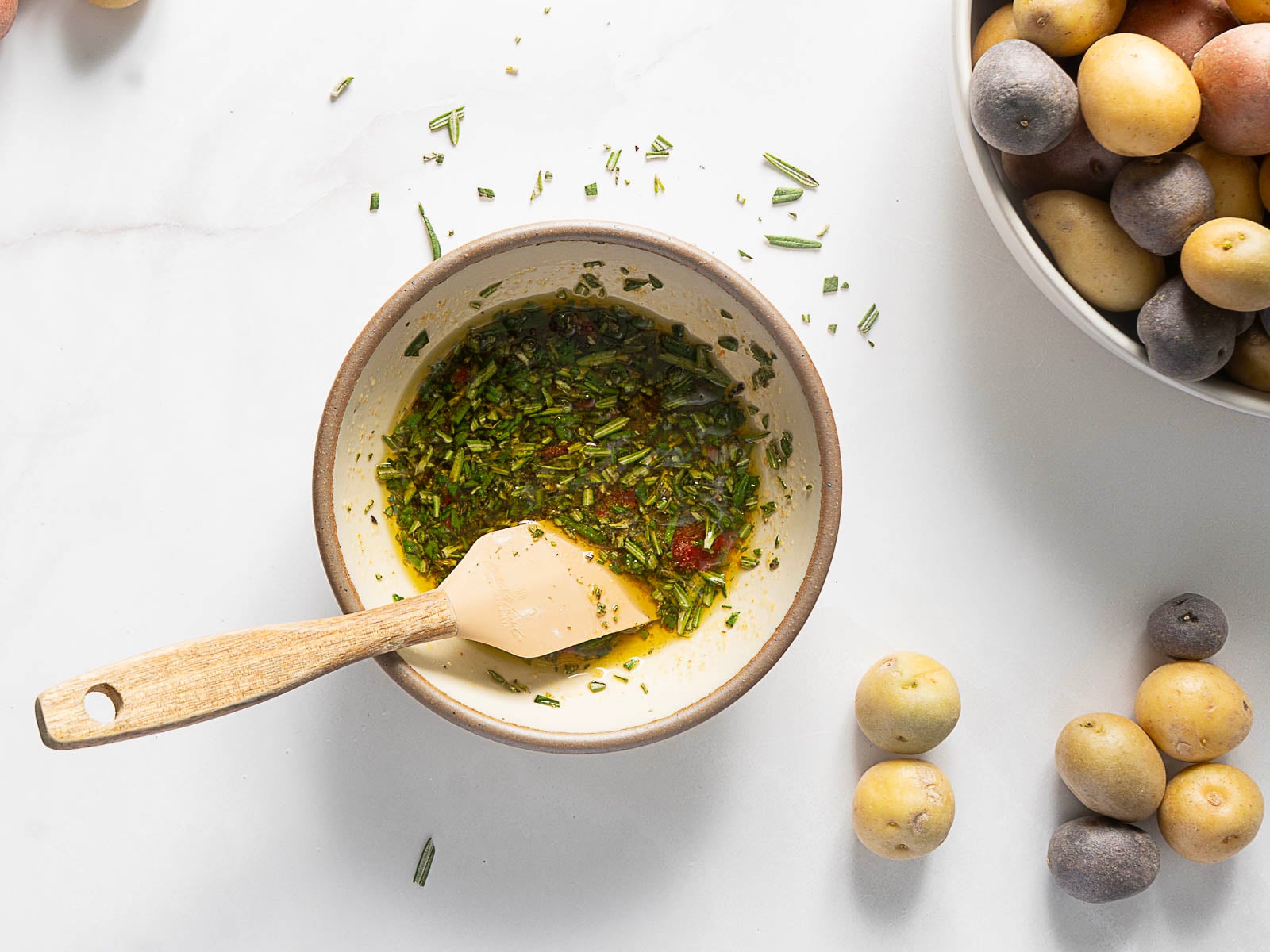 A bowl of herb and oil marinade with a spatula sits next to a bowl of assorted small potatoes, ready for making crispy baby potatoes on a white surface.