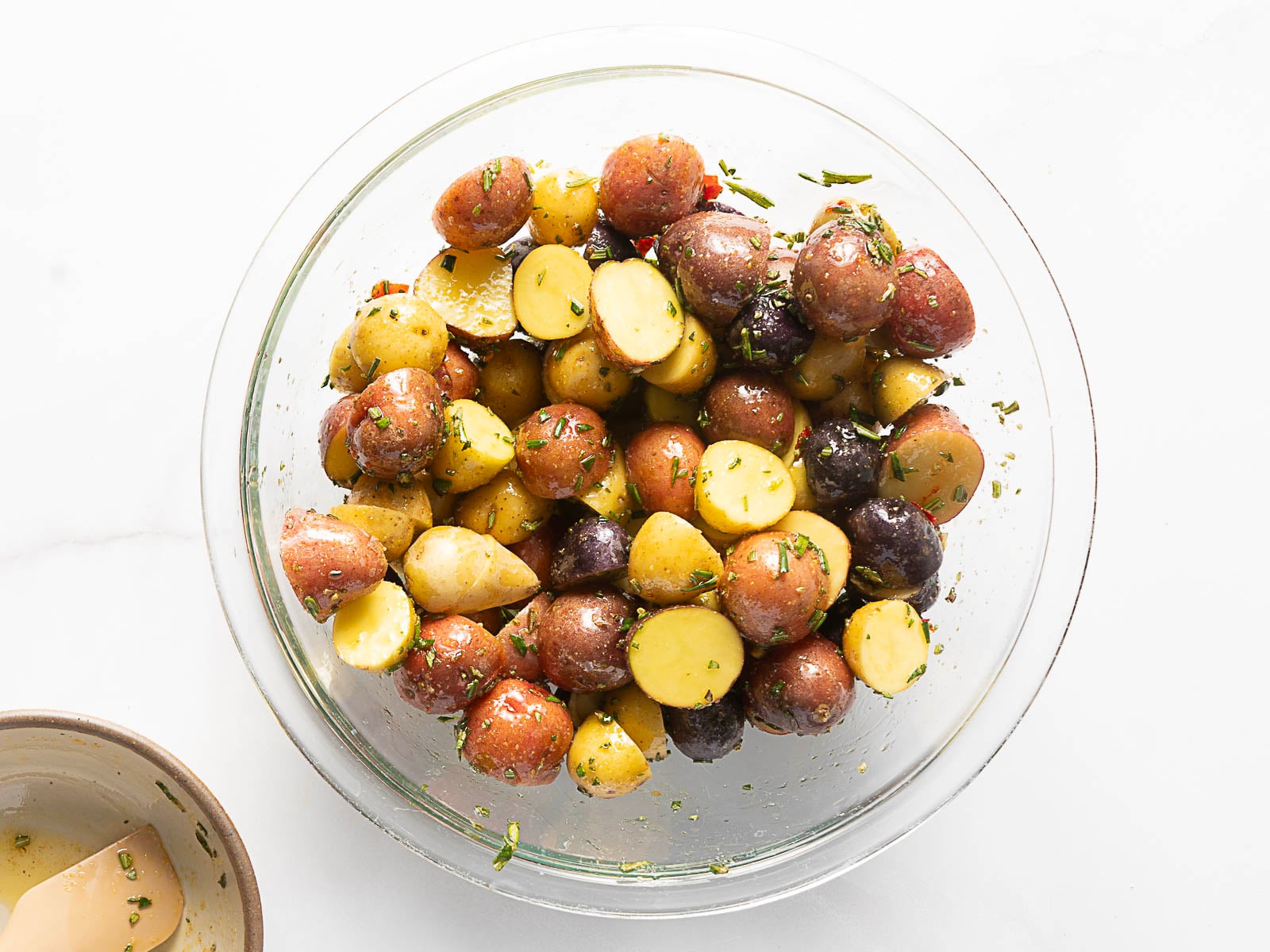 A glass bowl filled with halved and seasoned roasted baby potatoes sits on a white surface; a small dish with sauce is partially visible nearby.