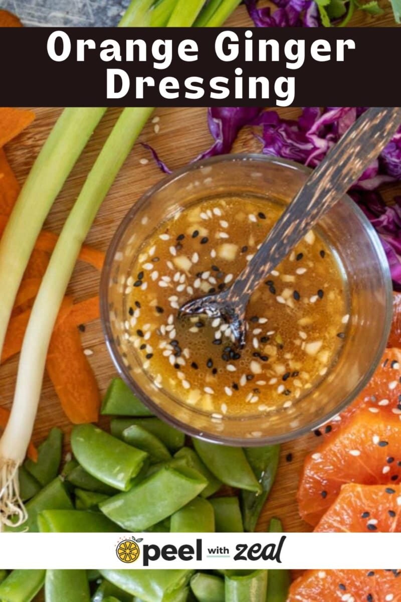 A bowl of orange ginger dressing with sesame seeds and a spoon, surrounded by fresh vegetables and orange slices on a wooden surface.